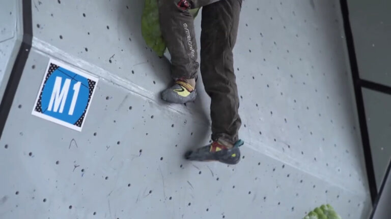 Close-up of a climber's feet wearing specialized climbing shoes on an indoor climbing wall marked with M1