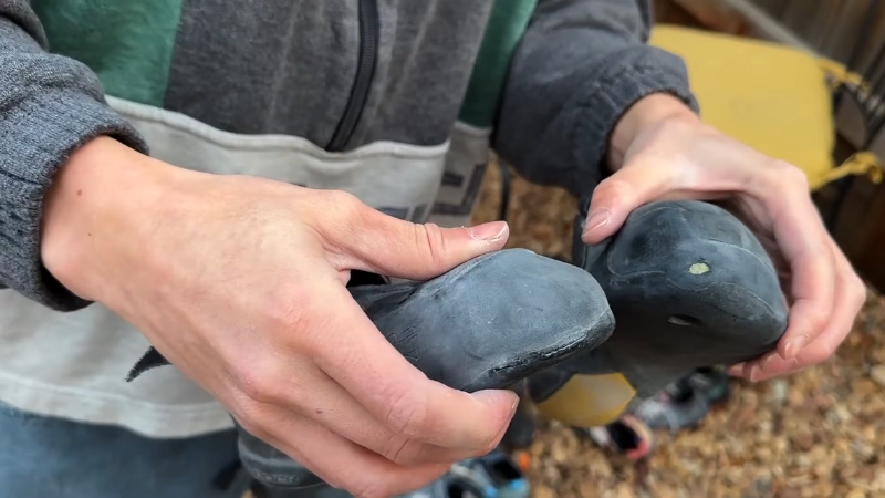 A climber holds a worn pair of bouldering shoes outdoors, showing signs of use and rubber wear