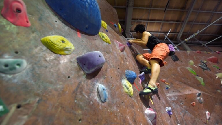 A climber in bright shorts moves across an indoor bouldering wall, focused on finding stable holds