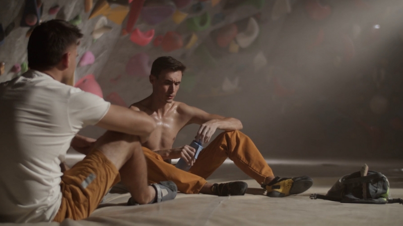 Two male climbers sit on the gym floor resting, one looking serious and focused after a climb