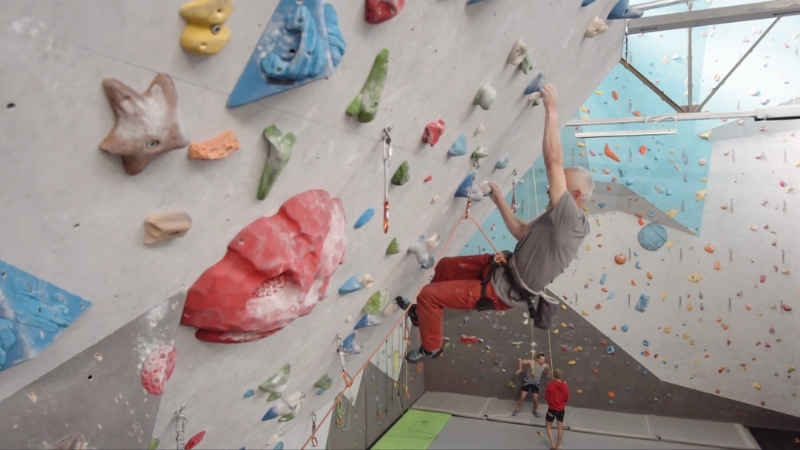 A man climbs a steep indoor wall, reaching for a hold while navigating a complex overhang route