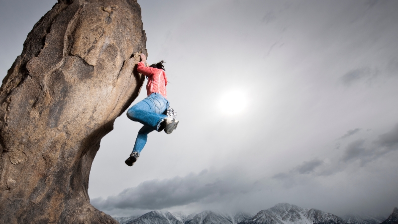 A climber hangs on a high rock face with snowy mountains and cloudy skies in the background