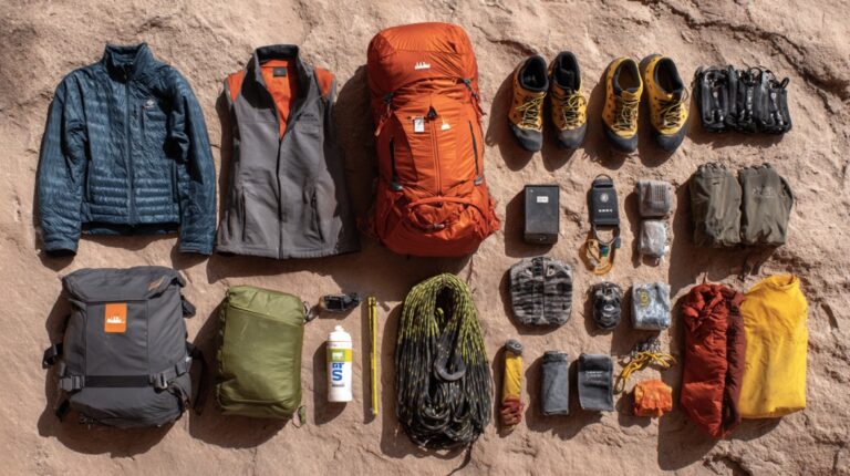 Flat lay of bouldering gear including backpack, climbing rope, shoes, jacket, vest, water bottle, sleeping bag, and other climbing essentials neatly arranged on rock surface