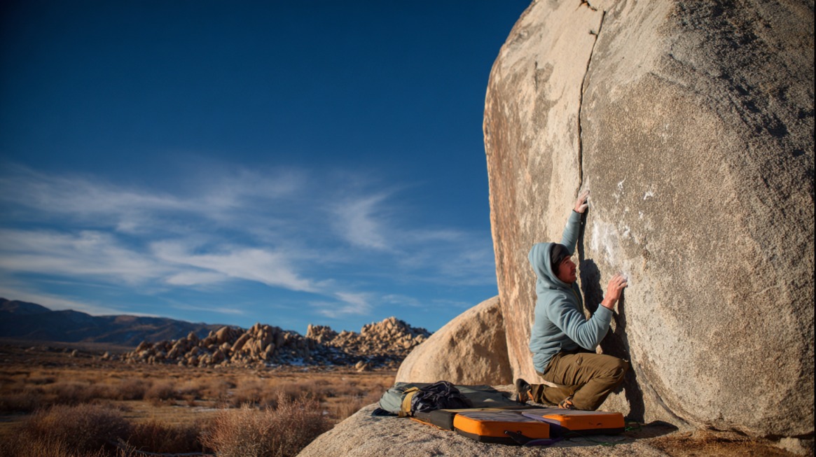 Climber in a hoodie starting up a large outdoor boulder with crash pads on the ground for safety