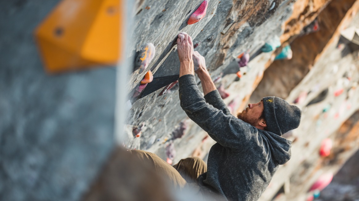 Man wearing a beanie and hoodie climbing a rock wall with colorful holds, focusing on grip and movement during a bouldering session